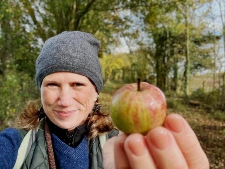 Lisa M. Rose in a grey beanie holding up a small wild apple in autumn woodland