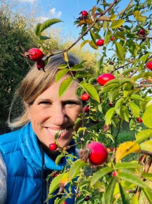 Lisa M. Rose smiling beside a wild rose bush heavy with red rose hips